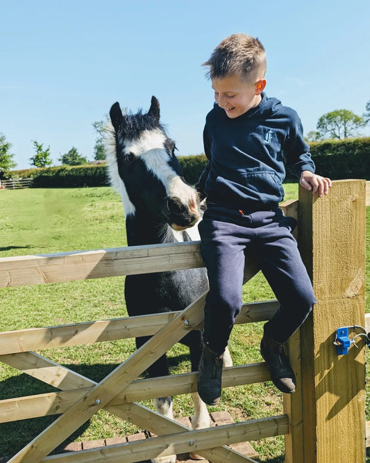 Young boy sitting on gate next to his piebald pony in the sunny weather